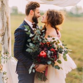 A bride and groom looking lovingly at each other, whilst holding hands and embracing a large bouquet of silvery green foliage, and red flowers with red berries